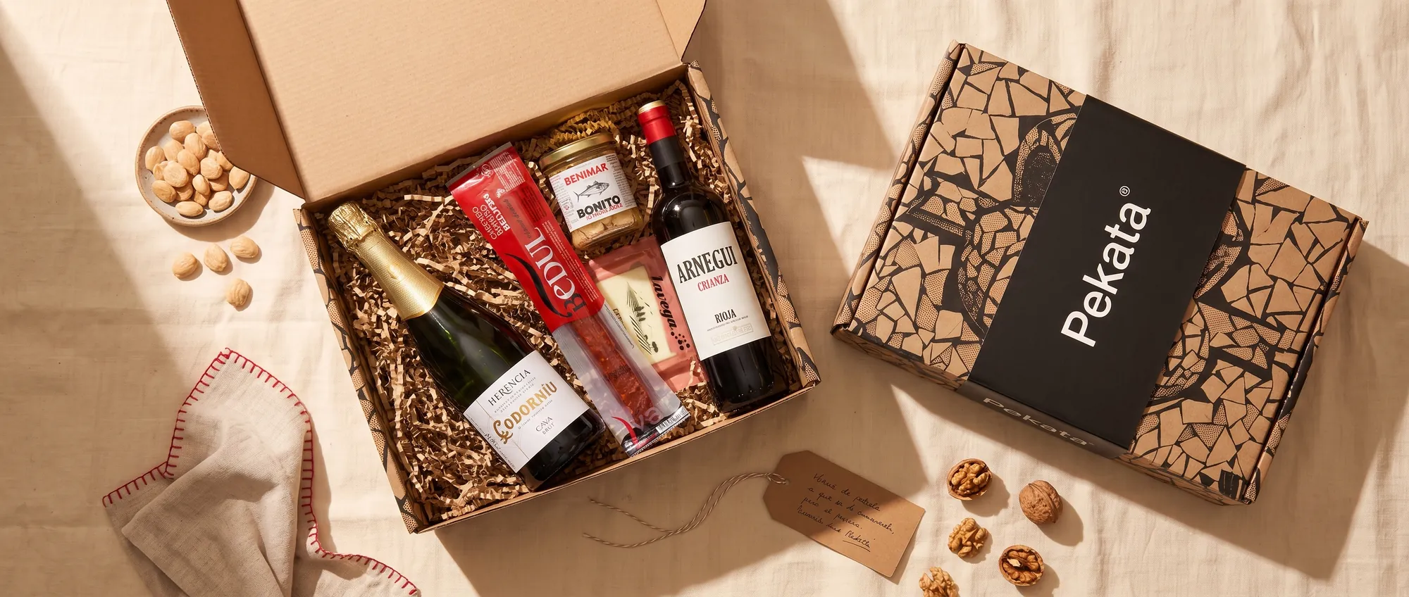 Caja Pekata abierta como cesta de Navidad, con cava, vino tinto, chorizo ibérico, bonito y queso curado, sobre un mantel de lino