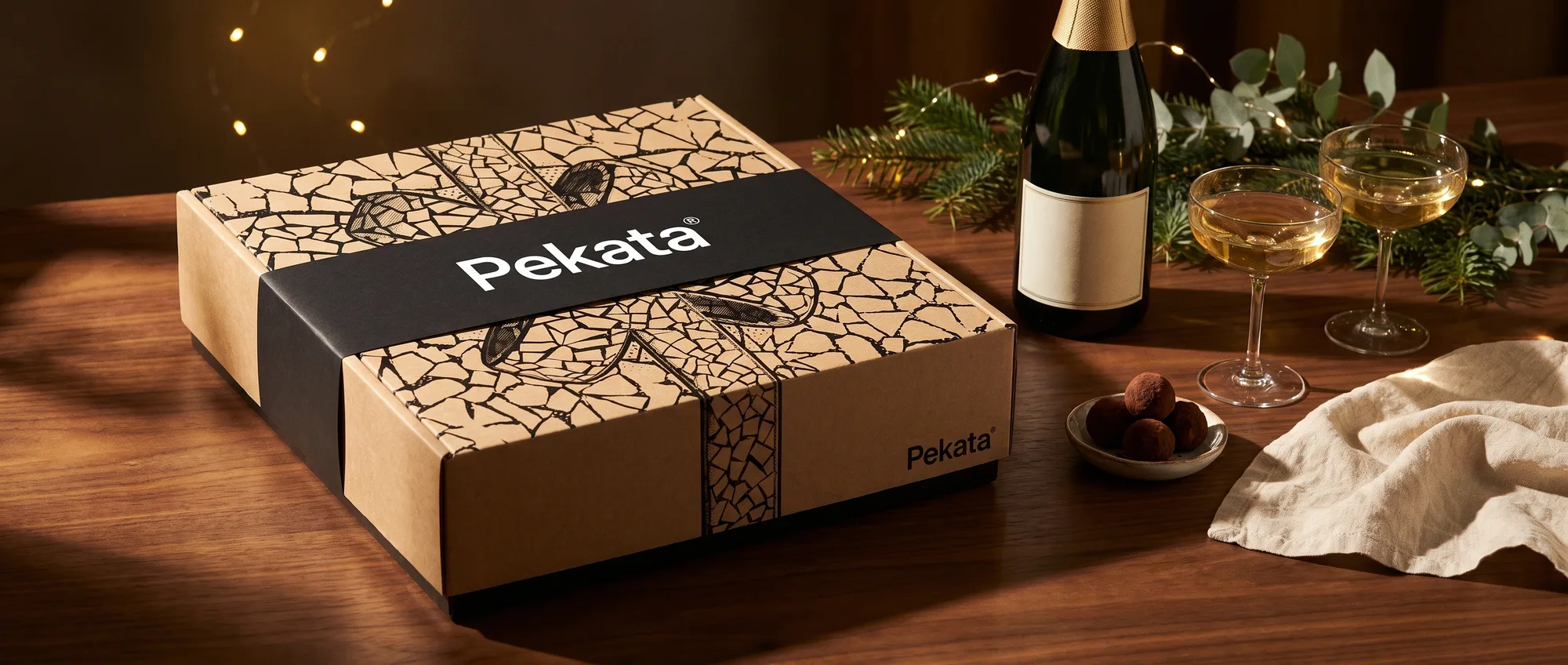 Caja de Navidad Pekata cerrada con faja negra junto a una botella de cava y dos copas sobre una mesa de nogal