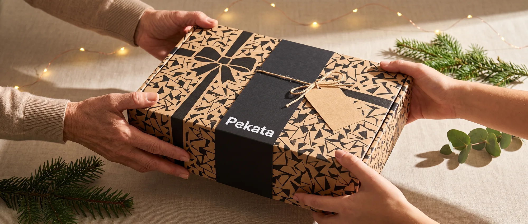 Dos pares de manos entregando y recibiendo una caja de regalo Pekata con etiqueta kraft y estampado trencadís